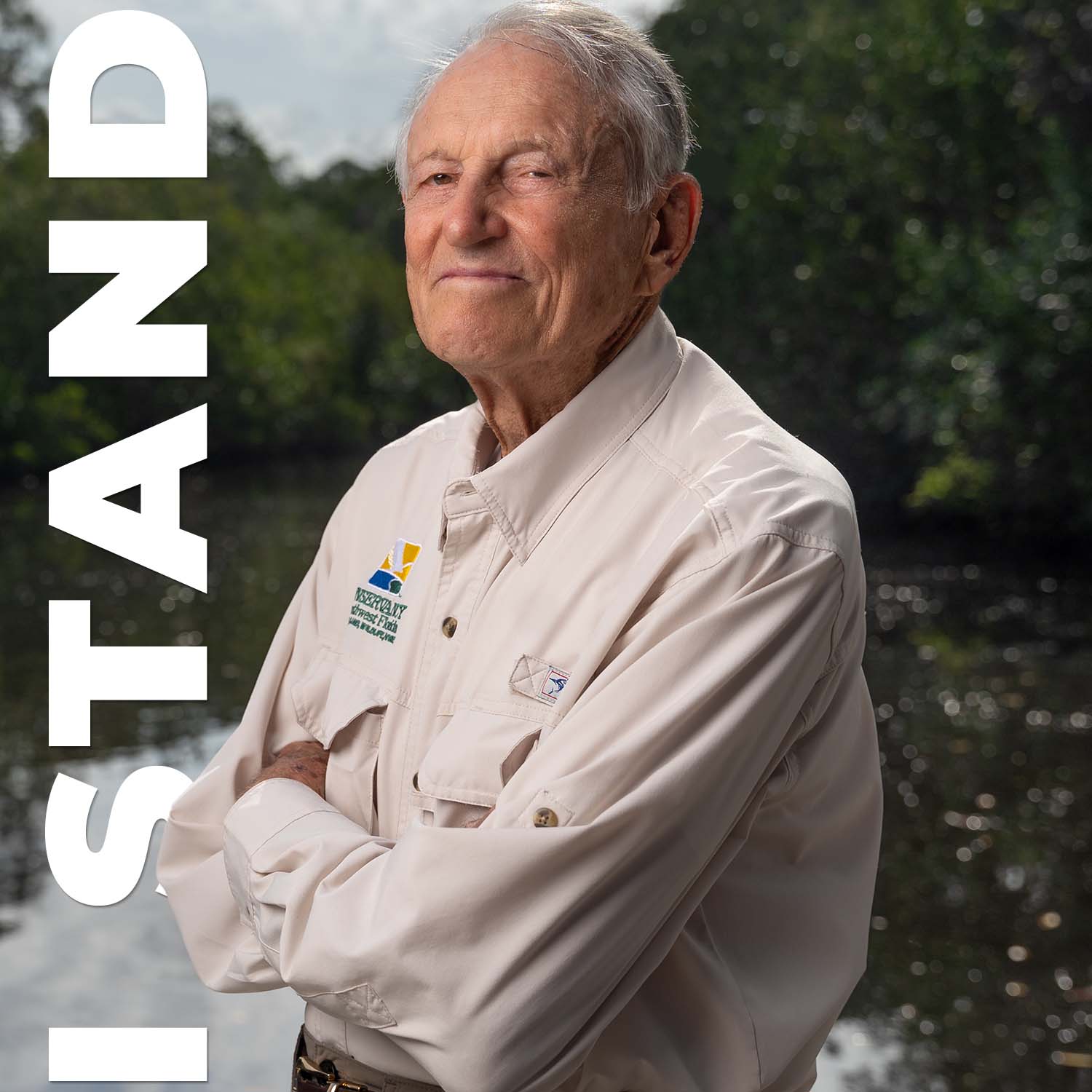 Senior male standing in front of a body of water with arms folded. Text along the side of image reads, "I Stand"