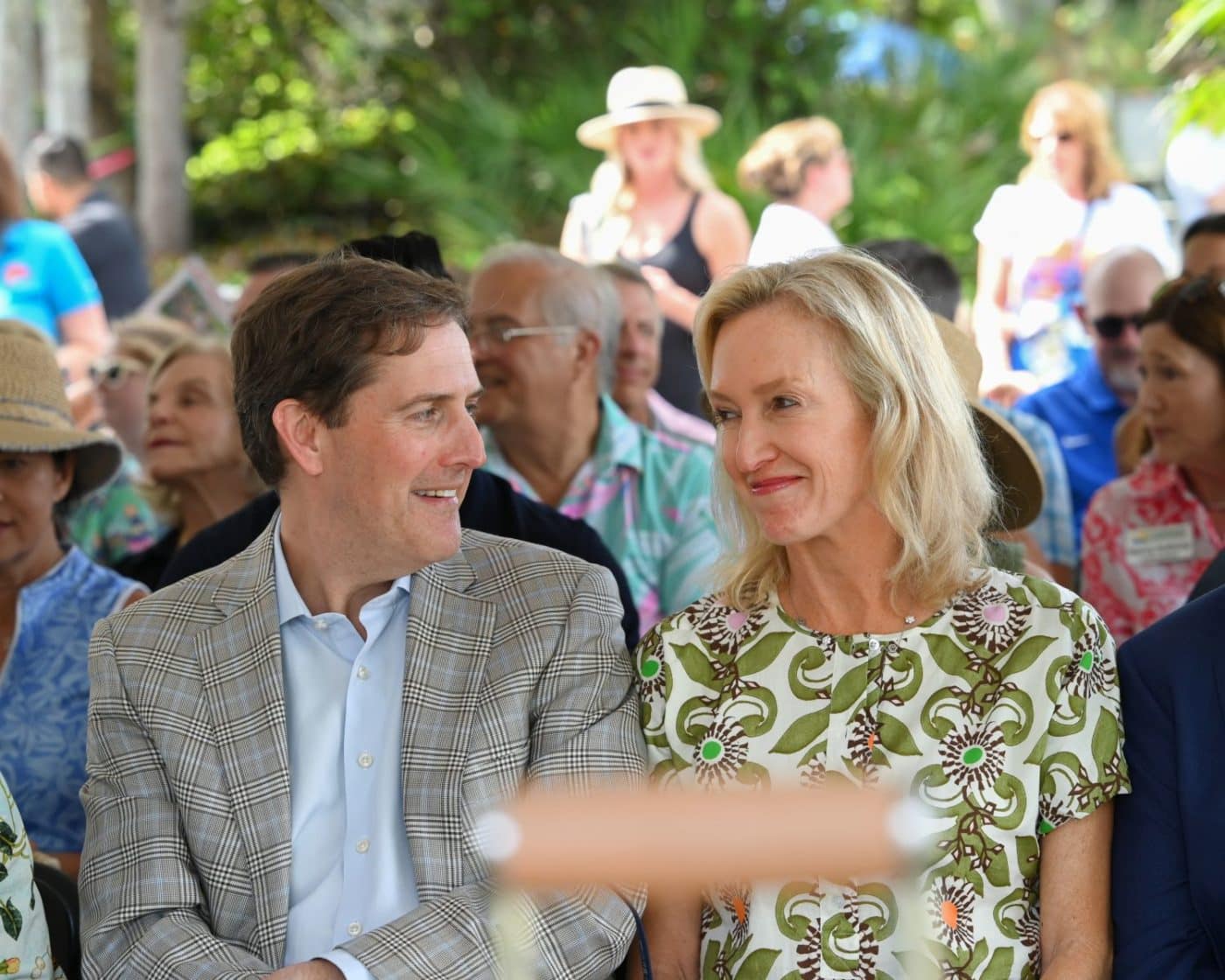 Man and woman seated outdoors at a social event, smiling at each other in the foreground.