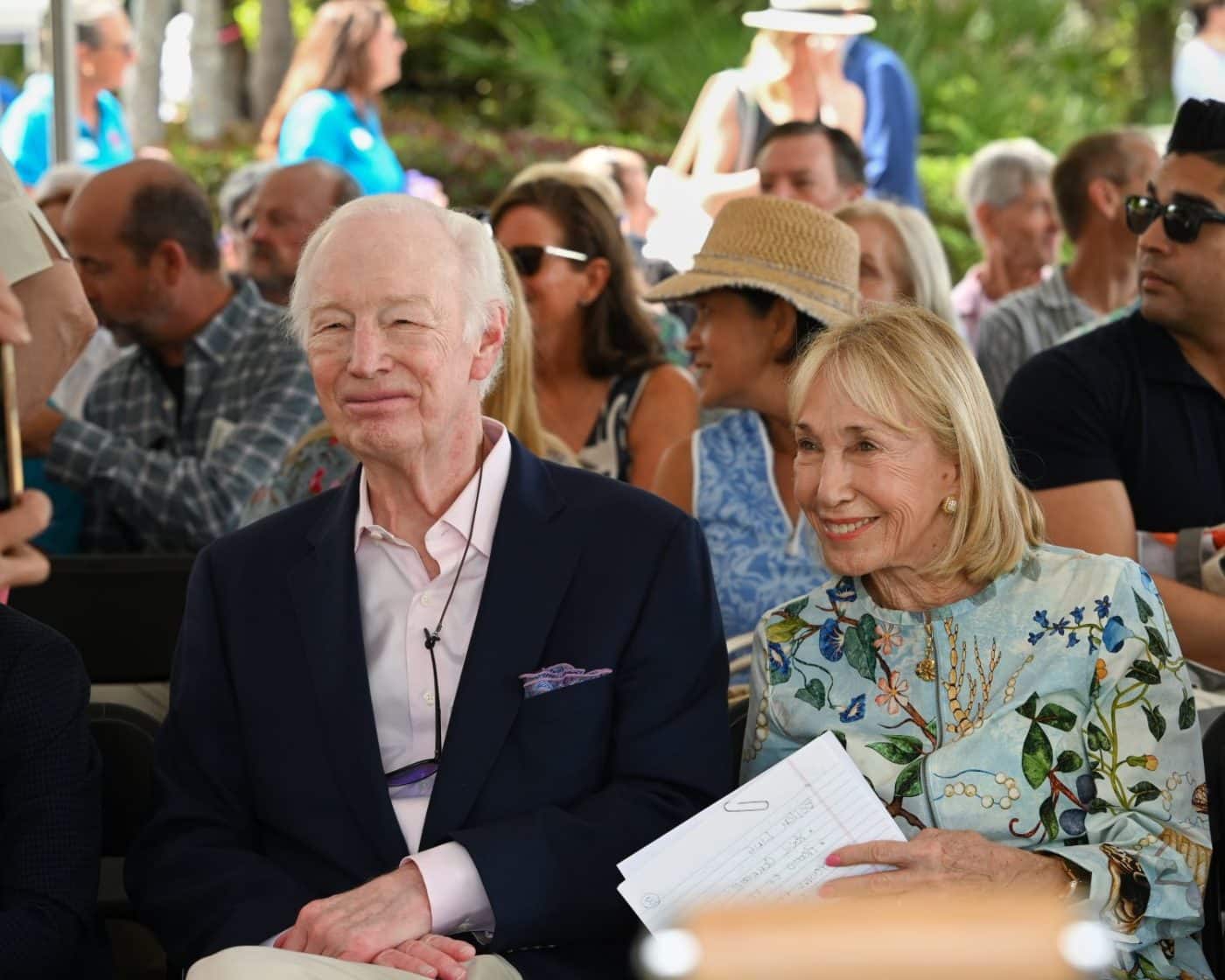 An elderly couple seated at an outdoor event, smiling; man in a navy blazer and pale pink shirt, woman in a floral blouse with papers in her hand.