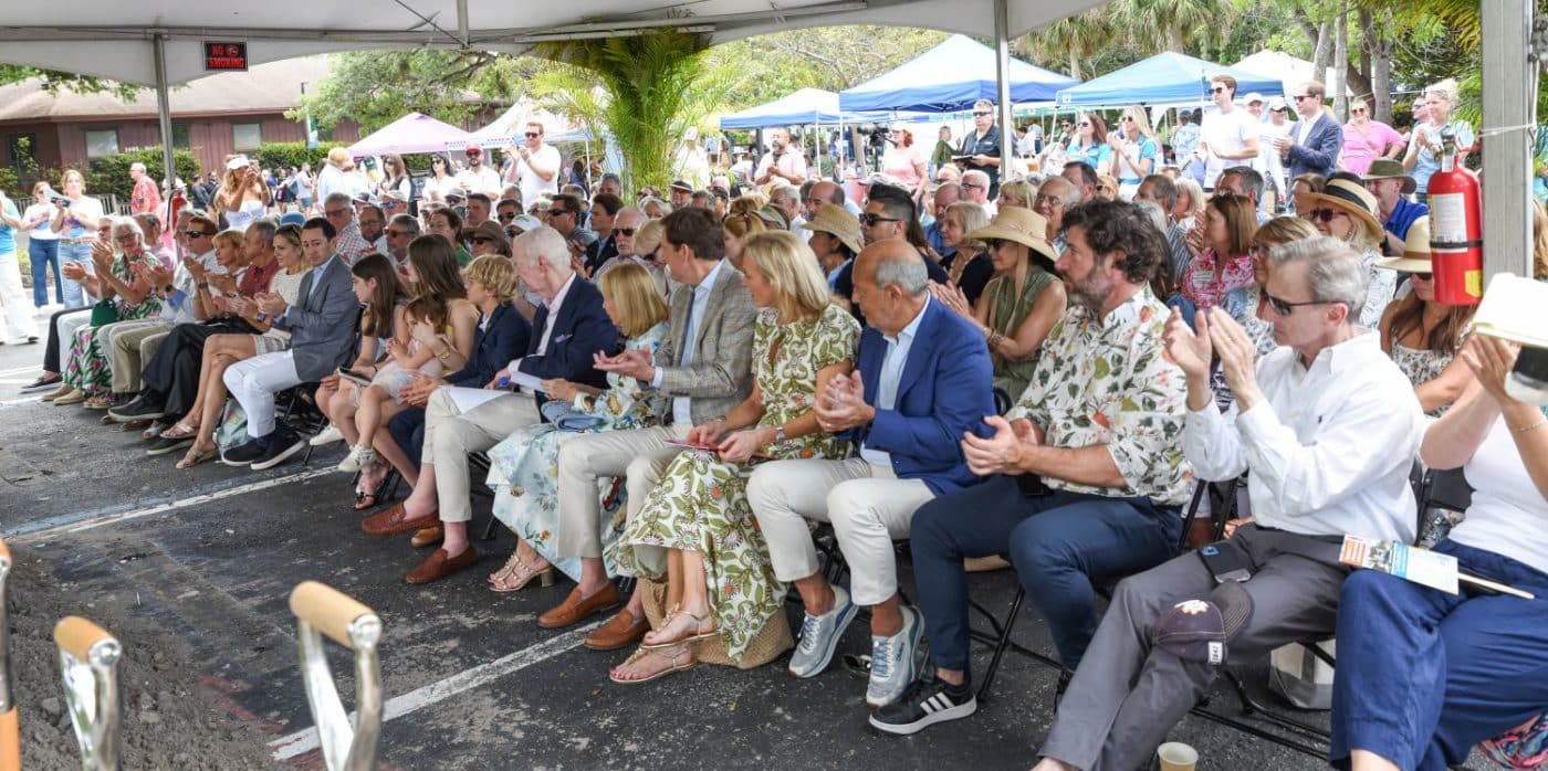 Crowd of people seated under a large canopy at an outdoor event, clapping; tents and onlookers in the background.