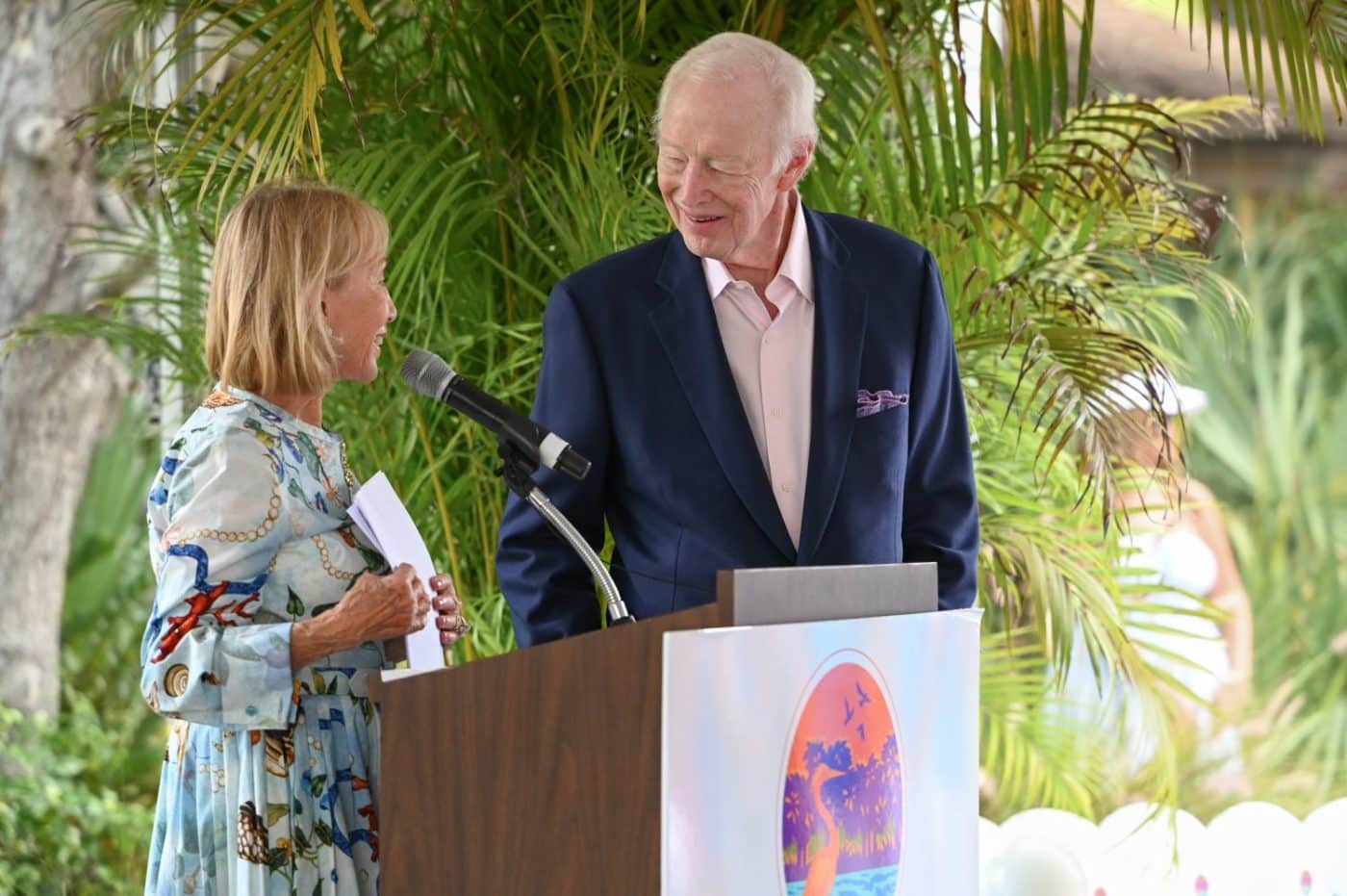 Two people speak at a podium outdoors, smiling at each other, with a microphone and tropical plants in the background.