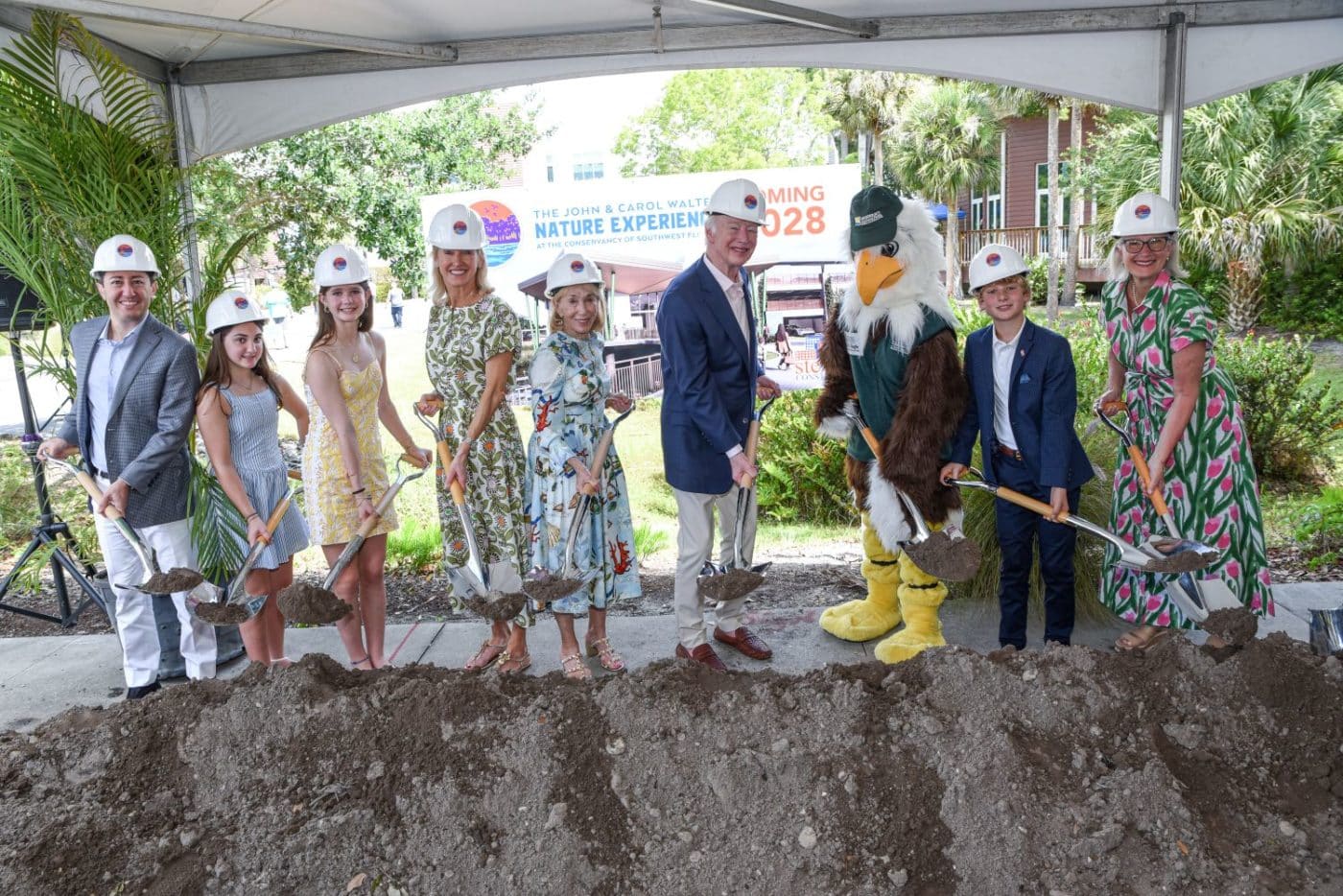 Groundbreaking ceremony with kids and adults in hard hats, holding shovels over a dirt mound under a tent, mascot nearby.